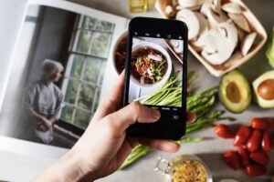 From above view of faceless blogger taking photo on smartphone of fresh tasty salad in white bowl and food placed around on table in modern kitchen